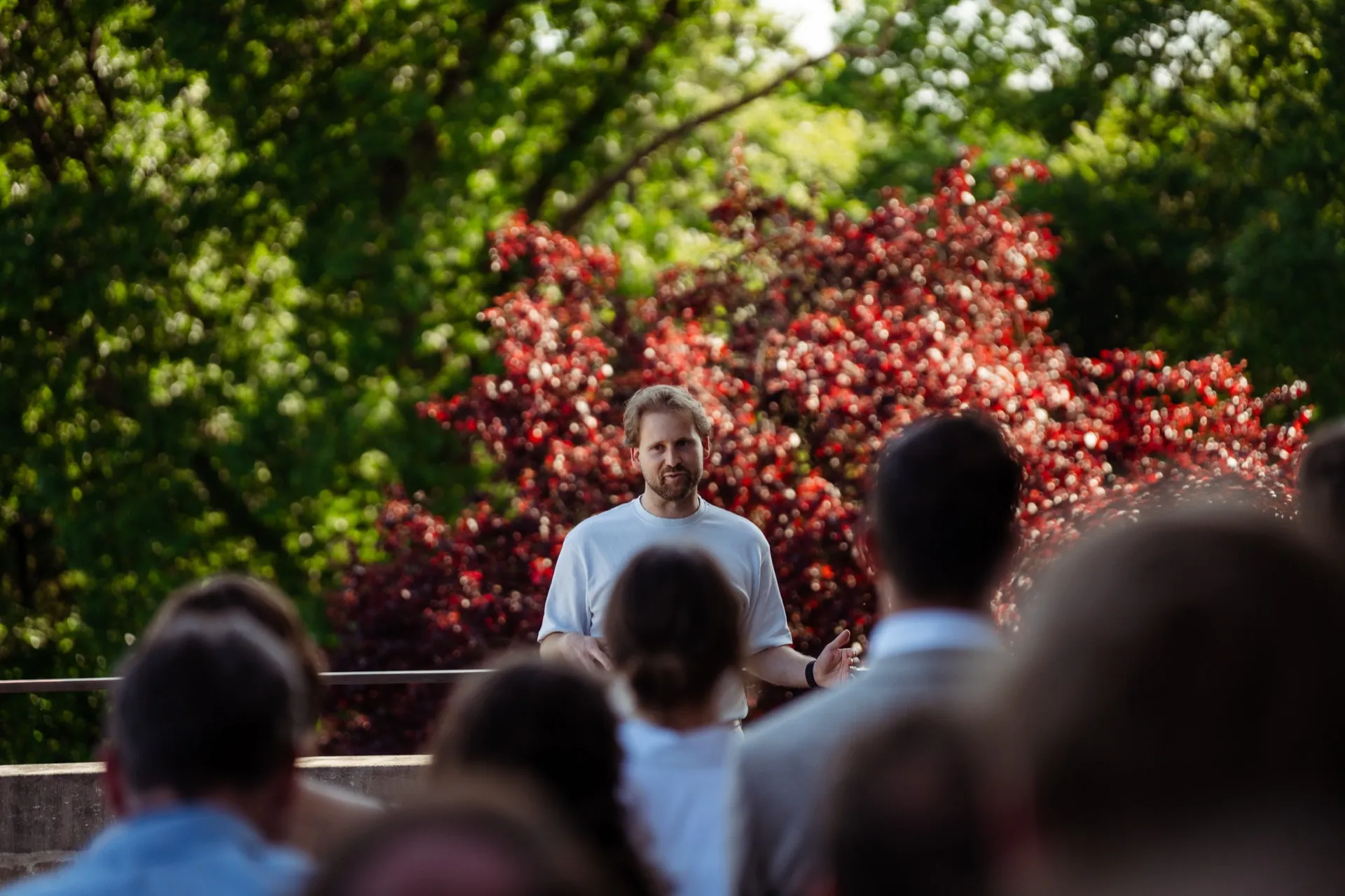 Warre Buysse spreekt een groep mensen toe in een tuin, voor een struik met rode bladeren.